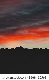 Vertical image of a dramatic sunset sky with deep red and dark gray clouds over a mountain silhouette. A moody and visually powerful scene ideal for posters, backgrounds, or emotional nature content.