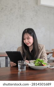 A vertical image featuring a young Asian woman looking down at a black digital tablet with a focused yet gentle smile. Dressed in a professional beige blazer, she sits at a wooden table in a minimalist dining area with a glass of water and a bowl of salad in front of her. Ideal for productivity mobile apps, workplace lifestyle photography, and healthy dining content.