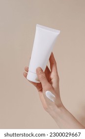 Vertical image of a female hand with a white smear holding a blank tube of moisturizing cream on a beige isolated background. Hand care, natural cosmetics and beauty concept. Image for your design - Shutterstock ID 2300029667