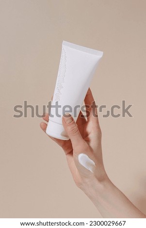 Vertical image of a female hand with a white smear holding a blank tube of moisturizing cream on a beige isolated background. Hand care, natural cosmetics and beauty concept. Image for your design