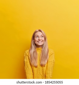 Vertical image of glad young European woman with blonde hair looks above happily has white teeth poses over yellow background empty space upwards for your promotion dressed in casual jumper.