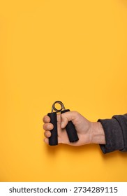 Vertical image of a hand using a grip strengthener isolated on a yellow background with copy space. Concept of hand strength training, physical rehabilitation, occupational therapy, stress relief, fitness, workplace wellness, and healthy lifestyle 