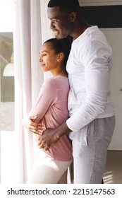 Vertical Image Of Happy Diverse Couple Embracing, Looking Out Of Window And Smiling At Home. Happiness, Domestic Life, Love And Inclusivity Concept.