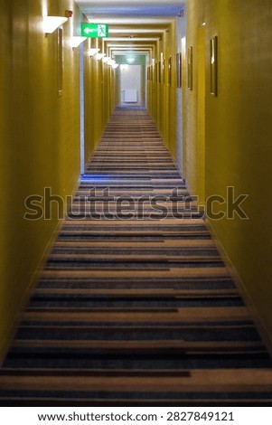 Vertical image of long corridor with yellow walls and green exit sign, strong perspective lines creating depth and direction in modern interior space.