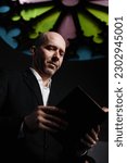 Vertical image of mature man in black suit reading Bible standing in old baptist church