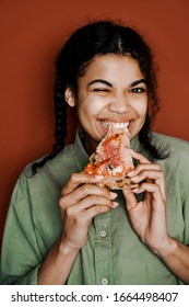Vertical image, overjoyed funny joyful smiling woman with pizza slice in hand. Happy young emotional teenager African American girl eating pizza isolated over orange studio background.