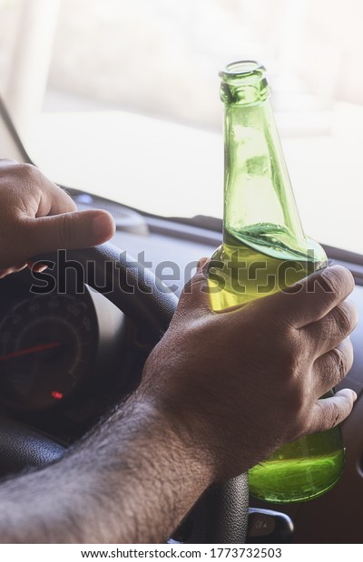 A vertical\
image of person driving a car while holding a bottle of beer. Drunk\
diving, unsafe driving\
concept