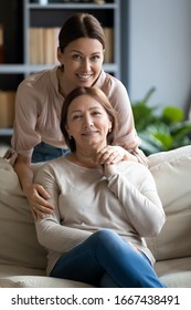 Vertical image portrait happy grownup daughter hugging pleasant older brunette mother resting on couch. Smiling young woman posing for bonding family photo with mom in living room, looking at camera.