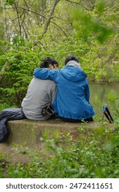 Vertical image of rear view of two people sitting on a rock near a river hugging