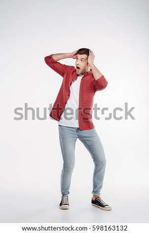 Vertical image of shocked man in shirt and jeans which holding head and looking away. Isolated white background