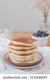 Vertical Image Of A Simple Traditional Stack Of Pancakes Maple Syrup Or Honey Running Down The Sides. Fruit, Flowers And Jug On Top A Wooden Table. Copy Space Available In Light Airy Backdrop