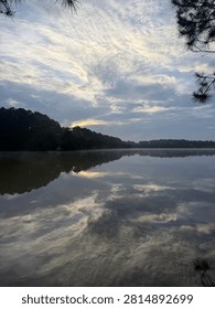 Vertical Image Of Sun Breaking Through Cloudy Sky Over Lake With Morning Mist Over Water And Tree Lined Horizon Outdoors