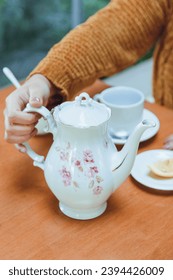 vertical image of tea time with unrecognizable woman in background and white porcelain teapot with cup and lemon slide on table.