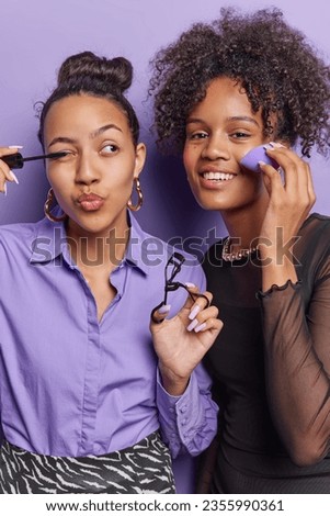 Vertical image of two young girfriends apply mascara put on makeup before going out get ready to night club stand closely to each other isolated over purple background. Women and beauty concept