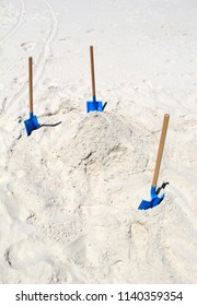 Vertical Image Of White Sandy Beach With Trio Of Bright Blue Shovels Ready To Help People Make A Sand Castle And Moat.