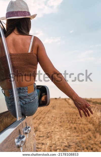 Vertical image of a woman sitting at the window\
of her camper van trying to capture the wind and the yellow wheat\
field with her hand. Van life\
concept.