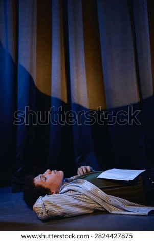 Vertical image of young man lying on stage floor with script on chest during student theater rehearsal, representing drama education and performing arts programs
