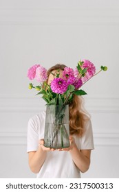 Vertical image of a young woman holding a bouquet of pink dahlias in a vase in front of her on a white background. The concept of flower workshops and articles for the study of floristry