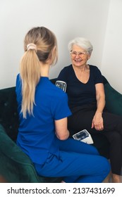 Vertical Indoor Shot Of Home Visit. Happy Senior Woman With Hyper Tension And Her Young Blonde Caregiver. High Quality Photo