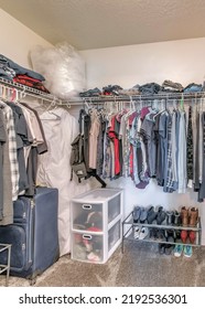 Vertical Interior Of A Small Walk-in Closet With Wall Mounted Wire Shelves. There Are Clothes Hanging Below The Shelves With Clothes And Boxes On Top And A Shoe Racks, Drawers And Bag