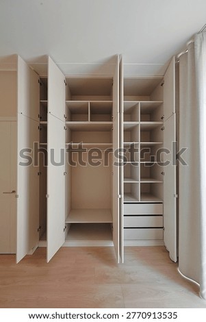 Vertical interior view of an open built-in wardrobe with shelves, hanging space, drawers, soft curtains, and light wood flooring in neutral tones