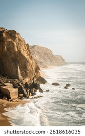 Vertical landscape of Atlantic ocean waves crashing against tall rocky cliffs on the West Coast of Portugal. Majestic rugged coastline illuminated by warm golden hour sunlight