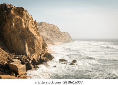 Vertical landscape of Atlantic ocean waves crashing against tall rocky cliffs on the West Coast of Portugal. Majestic rugged coastline illuminated by warm golden hour sunlight