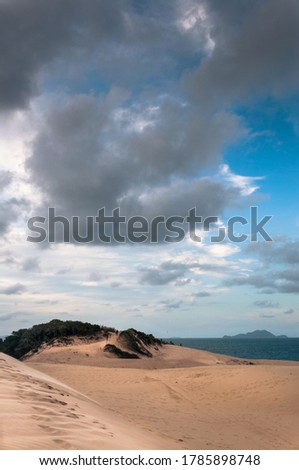 Vertical landscape of the dunes near the Praia dos Ingleses and Praia do Santinho, Florianopolis, Brazil. A big blue sky with white and gray clouds, the sea with a mountain in the background. 