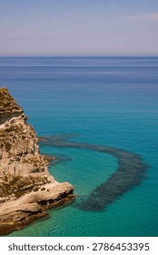 A vertical landscape view showcasing the dramatic coastal cliffs of Tropea in Calabria, Italy. The high-angle shot reveals crystal-clear, multi-toned turquoise water transitioning into a deep blue ocean horizon under a clear summer sky. Below the rocky sandstone promontory, a prominent curved underwater reef formation stands out starkly against the transparent sea floor. Perfect for mobile background layouts, travel guides, Mediterranean tourism marketing, and nature photography.
