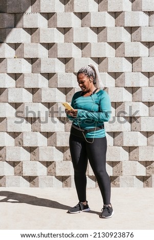 Vertical of Latina woman standing outdoors with cell phone in her hands getting ready to go for a run. 