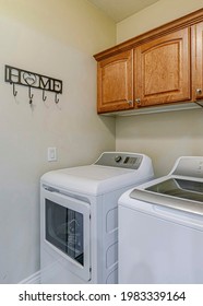 Vertical Laundry appliances inside small and functional utility room of residential home. Brown wooden hanging cabinet and hooks are mounted against the white wall.