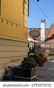 A vertical layout shot looking down a narrow cobblestone alleyway in a historic citadel, featuring a large blank oval metal sign hanging from a yellow building facade.