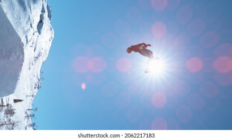 VERTICAL, LENS FLARE: Fearless Male Tourist Snowboarding In Fun Park Does A Big Backflip On A Perfect Winter Day. Breathtaking Shot Of Unrecognizable Man Snowboarding And Doing A Spectacular Stunt.