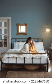 Vertical Lifestyle Photo Of Happy Young Mother And Daughter. Full Body Tender Mom And Baby Girl Sitting On Bed And Hugging At Cozy Bedroom With Modern Retro Interior. Happy Mothers Day. 