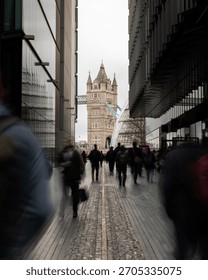 A vertical long exposure shot capturing Tower Bridge in London, UK, framed by modern glass office buildings, with blurred commuters walking rapidly toward the historic landmark.