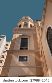 Vertical low angle looking up view of a traditional church bell tower and clock tower with a cream and peach colored facade against a clear blue sky.