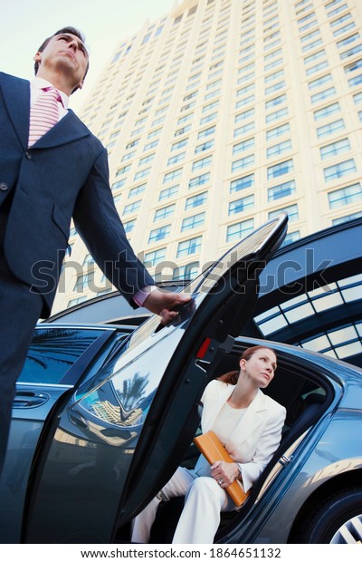 Vertical low\
angle shot of a chauffeur opening the car door for a businesswoman\
outside the office\
building.