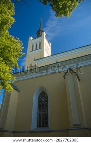 Vertical low angle shot of the neogothic St. John's Church in Tallinn, Estonia. Pastel yellow facade with bell tower, green tree leaves, harsh shadows and bright blue sky