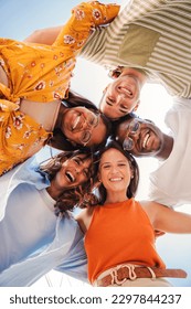 Vertical. Low angle view of a group of five multiracial teenagers smiling and looking down at camera outdoors. Young student people of diverse ethnic cultures embracing while standing in a circle