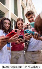 Vertical. Low angle view of group of Caucasian young people looking smiling videos on cell phone outdoors. Three generation z students friends having fun standing watching mobile together on campus