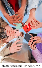 Vertical low angle view of a group of unrecognizable multiracial young friends using their cellphones on a circle. Team of teenagers enjoying and browsing on internet with a smart phone application