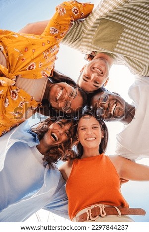 Vertical. Low angle view of a group of five multiracial teenagers smiling and looking down at camera outdoors. Young student people of diverse ethnic cultures embracing while standing in a circle