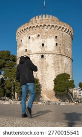 Vertical low angle view of a man in winter jacket walking towards the White Tower in Thessaloniki. Male tourist seen from behind adjusting his hair while exploring the city waterfront in Greece