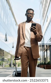 Vertical low angle of young Black man walking along city street and using smartphone while being on business trip