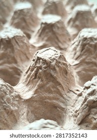 A vertical low-angle macro photograph capturing a prominent conical peak of an empty recycled paper pulp egg tray in sharp focus. The shallow depth of field causes the surrounding pulp cones and cavities to fade into a smooth, creamy background bokeh blur, creating a dynamic sense of depth, scale, and artistic perspective. The rough pressed fiber texture, crinkled surface, and natural light tan coloring of the central peak are illuminated by soft directional light, showcasing the sturdy yet lightweight cushioning design of the eco-friendly container. This creative visual composition is ideal for sustainable material research covers, zero-waste marketing campaigns, environmental packaging articles, agricultural supply blog graphics, circular economy concept banners, and abstract industrial stock photography celebrating biodegradable product innovations. The unique perspective emphasizes the structural integrity and eco-conscious craftsmanship found in everyday recycled cardboard packaging solutions.