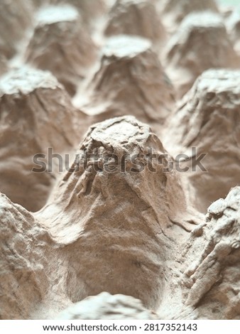 A vertical low-angle macro photograph capturing a prominent conical peak of an empty recycled paper pulp egg tray in sharp focus. The shallow depth of field causes the surrounding pulp cones and cavities to fade into a smooth, creamy background bokeh blur, creating a dynamic sense of depth, scale, and artistic perspective. The rough pressed fiber texture, crinkled surface, and natural light tan coloring of the central peak are illuminated by soft directional light, showcasing the sturdy yet lightweight cushioning design of the eco-friendly container. This creative visual composition is ideal for sustainable material research covers, zero-waste marketing campaigns, environmental packaging articles, agricultural supply blog graphics, circular economy concept banners, and abstract industrial stock photography celebrating biodegradable product innovations. The unique perspective emphasizes the structural integrity and eco-conscious craftsmanship found in everyday recycled cardboard packaging solutions.
