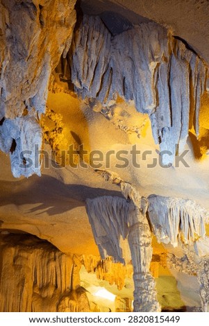 A vertical low-angle shot captures heavy, fluted stalactites and layered limestone curtains hanging from a cavern ceiling illuminated by contrasting warm and cool artificial lighting inside a cave in Ha Long Bay, Vietnam.