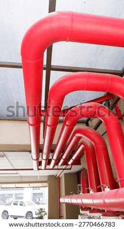 A vertical low-angle shot of large red steel pipes used for a building's fire suppression system. The pipes feature 90-degree elbows curving upwards toward a translucent polycarbonate roof, showcasing industrial plumbing installation and fire safety infrastructure.