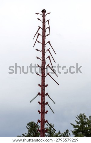 A vertical, low-angle shot of a tall, rustic wooden totem or memorial pole adorned with numerous antique mining pickaxes and axes pointing outward at various angles. The weather-beaten wooden post stands tall against a flat, white overcast sky, with the dark green tips of pine trees visible at the base of the frame. This poignant structural monument serves as a historical tribute, with each individual pickaxe representing a miner who tragically lost their life working underground in the local iron mines.