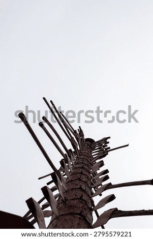 A vertical, low-angle shot of a tall, rustic wooden totem or memorial pole adorned with numerous antique mining pickaxes and axes pointing outward at various angles. The weather-beaten wooden post stands tall against a flat, white overcast sky, with the dark green tips of pine trees visible at the base of the frame. This poignant structural monument serves as a historical tribute, with each individual pickaxe representing a miner who tragically lost their life working underground in the local iron mines.